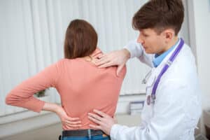 Doctor examines a woman’s back, palpating the spine while she stands with hands on her hips, suggesting a back pain assessment